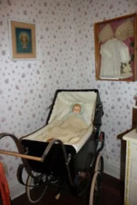 An upstairs bedroom in the sheriff’s residence with patterned wallpaper, framed portraits, and an antique wicker baby carriage.