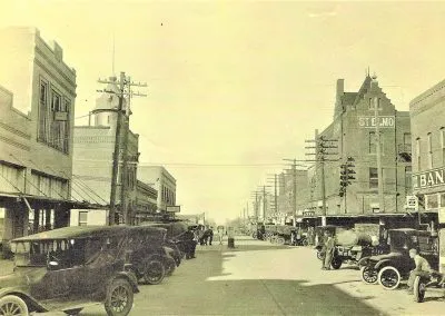 A sepia view down Bridge Street: brick buildings with awnings and early automobiles parked along the curb.