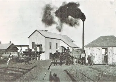 Cotton-gin buildings with a smokestack trailing smoke and wagons in the yard.