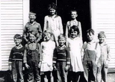 Schoolchildren lined up on the steps of a white frame schoolhouse.
