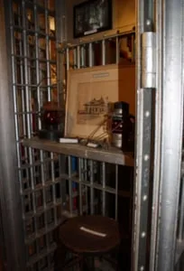 A jail cell seen through its open barred door: a narrow bunk with a blanket, a small window, and riveted steel walls.