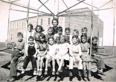 Young children and their teacher posed in rows outside a school.