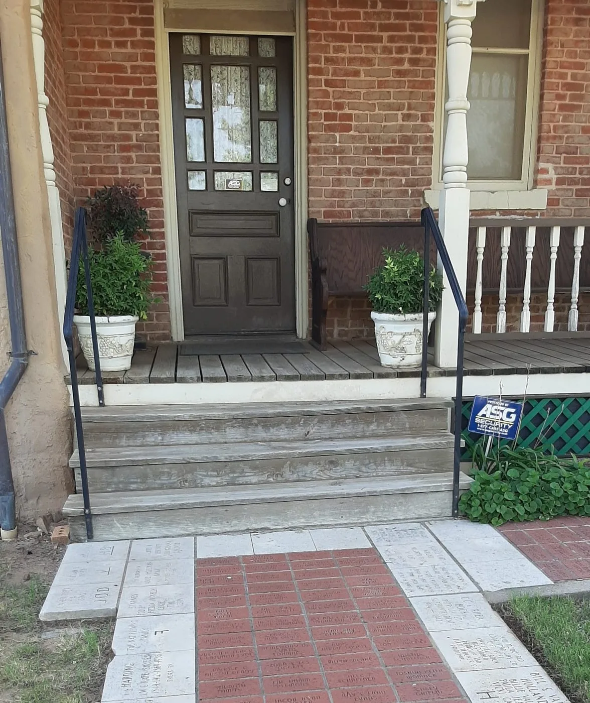The museum porch with its dark front door and potted plants, reached by a red-brick walk engraved with donors’ names.