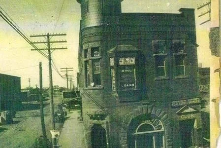 A hand-tinted photograph of an ornate two-story brick-and-stone building with a corner tower and tall arched windows.