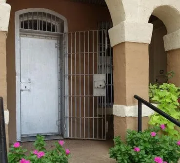 The arched front entrance of the jail wing: a heavy steel door with a barred window, set inside a stucco arch, with flowering plants at the foot of the steps.