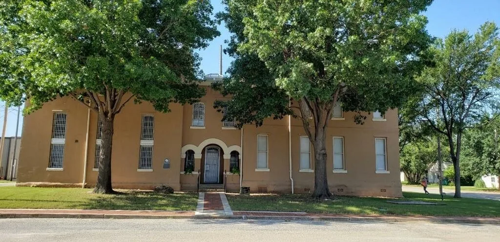 The jail wing of the 1890 Clay County Jail: a two-story tan stucco building with tall barred windows, a Roman-arched entrance, and a Texas Historical Commission marker beside the door, seen from the street through two large shade trees.