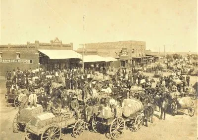 Wagons piled with cotton line a dirt main street in front of frame storefronts.
