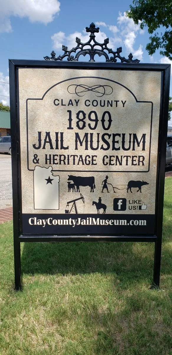 The museum’s roadside sign in a black iron frame: “Clay County 1890 Jail Museum & Heritage Center,” with silhouettes of a county map, longhorn, plow, oil derrick, and cowboy.