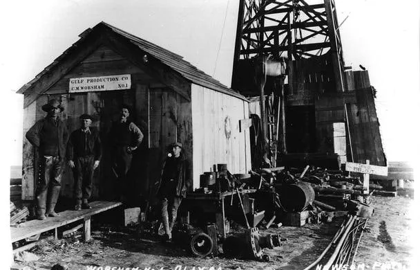 A black-and-white photograph of four oil-field workers beside a wooden shack and a drilling derrick.