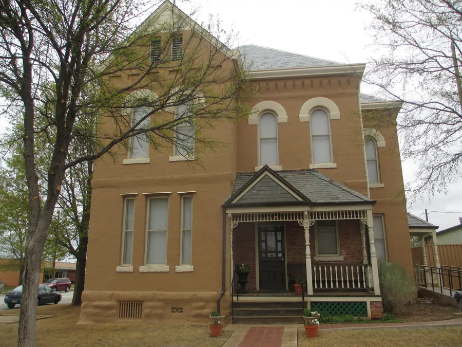The residence wing of the 1890 Clay County Jail: a two-story tan stucco house with a steep gable, Roman-arched upper windows, a decorative brick cornice, and a small gingerbread-trimmed front porch with potted plants on the steps.