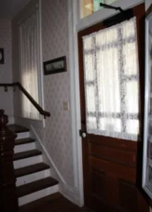 Inside the sheriff’s residence: a wooden staircase beside the front door, whose window is hung with a lace curtain.