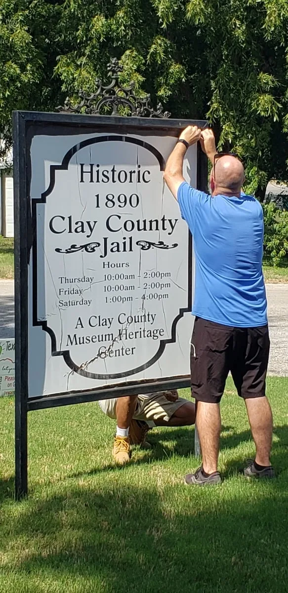 A volunteer in a blue shirt reaches up to lift the old “Historic 1890 Clay County Jail” panel out of its frame.