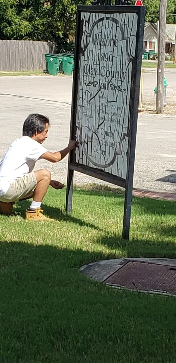 A volunteer crouches at the base of the sign frame, scraping the old sign panel.