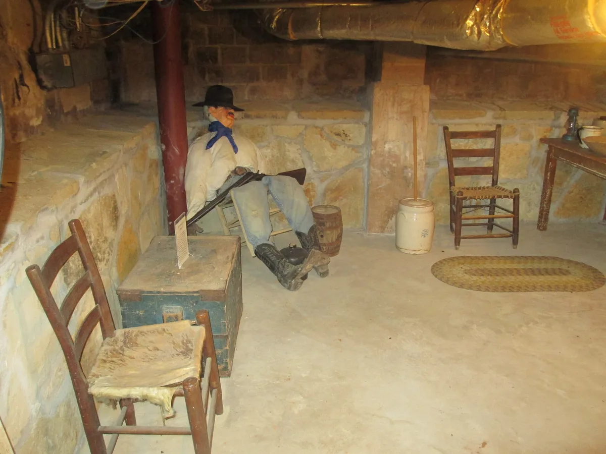 The sleeping guard exhibit in the stone-walled basement: a figure in a cowboy hat dozes on a wooden chair with a rifle across his lap, beside an old trunk and a second empty chair.