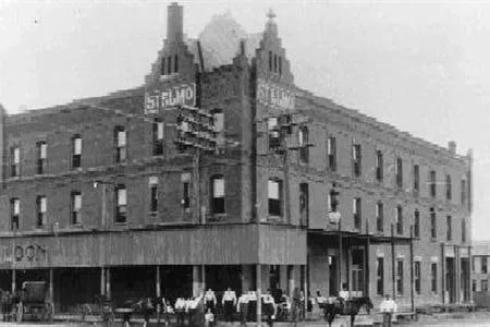 A three-story brick hotel with a crowd of people gathered on the sidewalk in front of it.