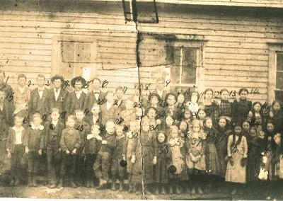 A large group of students in rows in front of a frame schoolhouse.