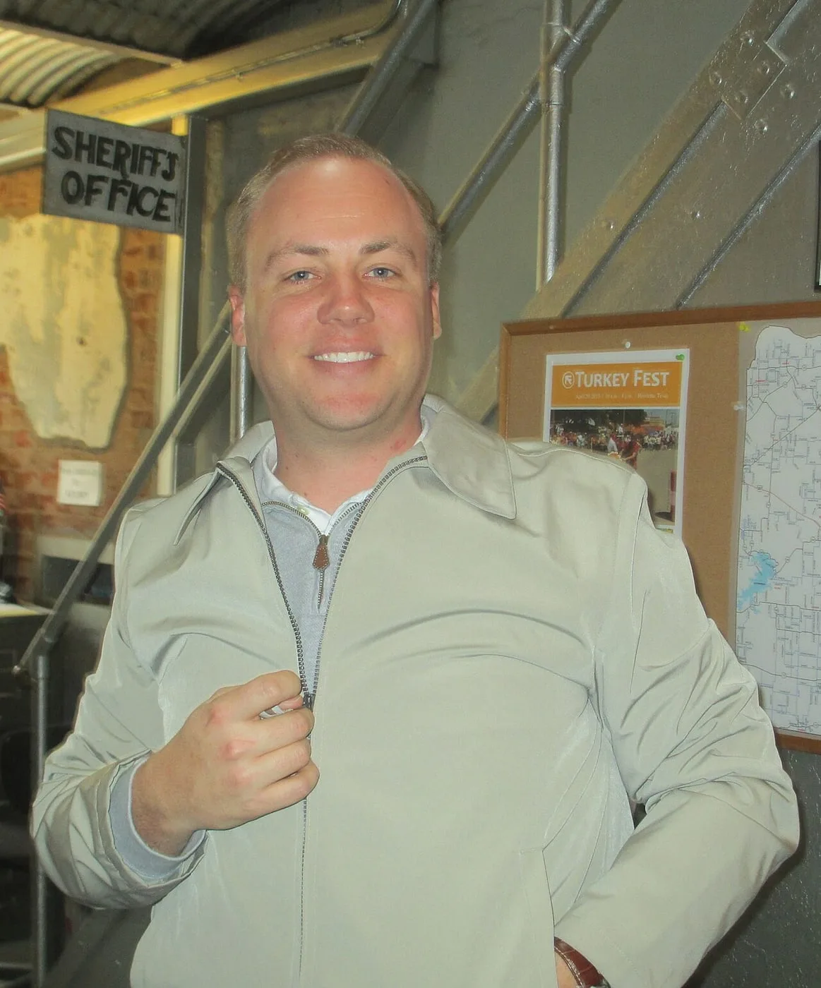 A smiling man in a light jacket stands inside the museum beside a wall of framed displays, under a sign reading “Sheriff’s Office.”